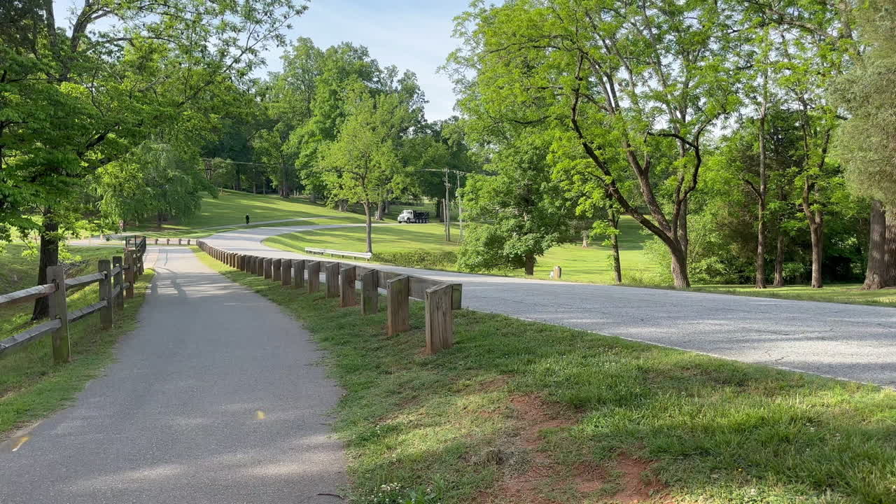 A quiet tree-lined walking path with a wooden fence winds through a lush green park