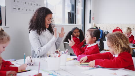 Young-female-infant-school-teacher-sitting-at-a-table-in-a-class-with-her-pupils,-counting-with-girl