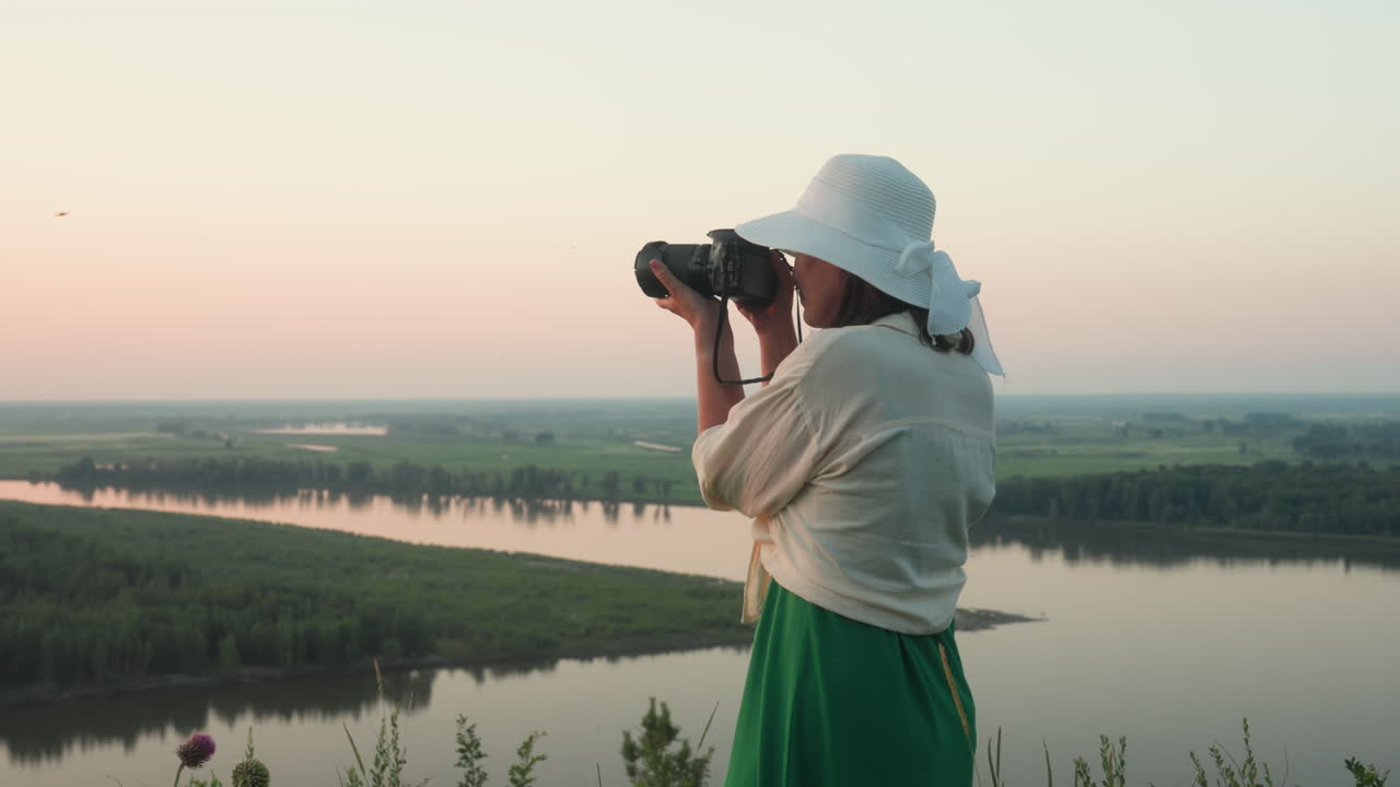 side view of woman in white sun hat holding DSLR camera while photographing scenic river valley at sunset from hilltop vantage point under soft pastel evening sky