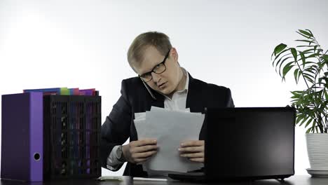 a man in a suit and glasses is sitting at his desk in an office, talking on the phone and looking at some papers. he has a laptop and a plant behind him. the man is focused on the conversation and appears to be busy with his work.