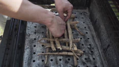 arms of a white caucasian man placing small wooden sticks on a barbecue surface with his hands