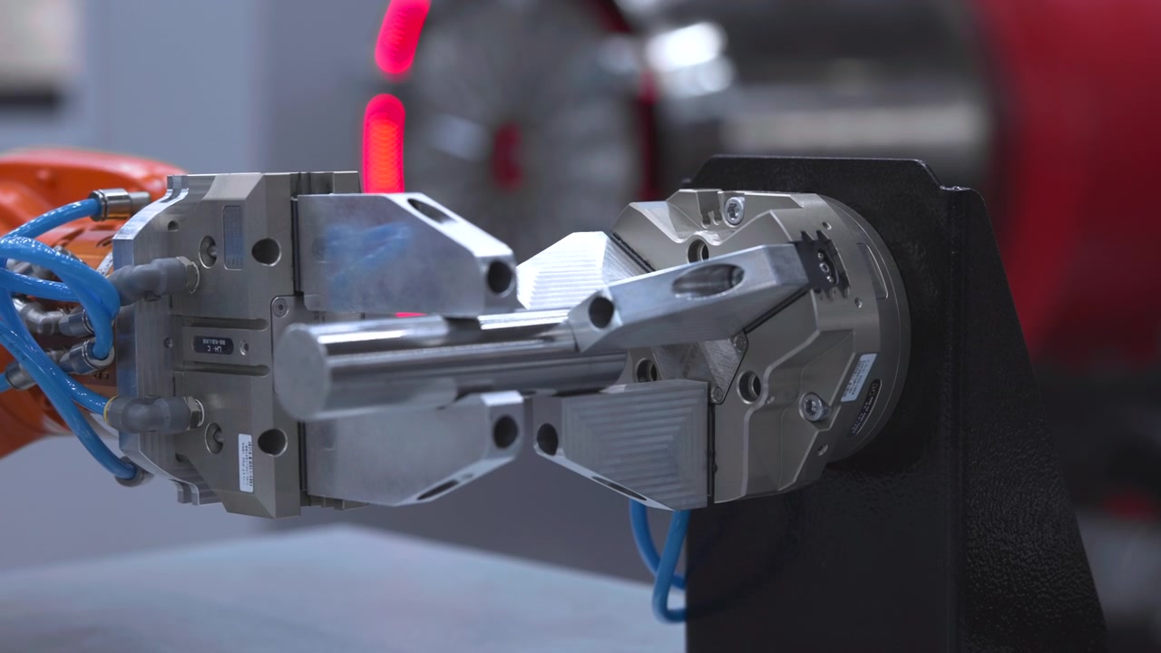 産業用ロボットが円筒形の金属棒を回転するチャックジョーに配置し、部品が自動加工セル内でボルトや青いチューブと共に正確にクランプされる様子を接写で捉えたもの。