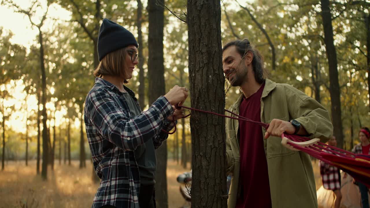 A happy girl in a black hat in a plaid shirt with glasses is tying up one side of a hammock, she is helped in this by a brunette man in a light green jacket during a hike in the summer forest