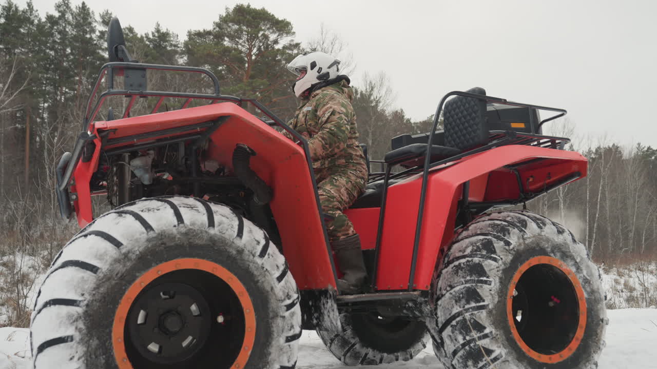 ATV rosso, foresta innevata, pilota mimetico che naviga un sentiero ghiacciato, pilota con casco ispeziona la macchina con pneumatici da neve sovradimensionati, cerchi arancioni, brina sulla carrozzeria, sfondo di pini e betulle, cielo coperto, alito freddo, ambiente impervio.