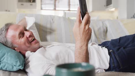 happy caucasian man lying on sofa in living room, using smartphone