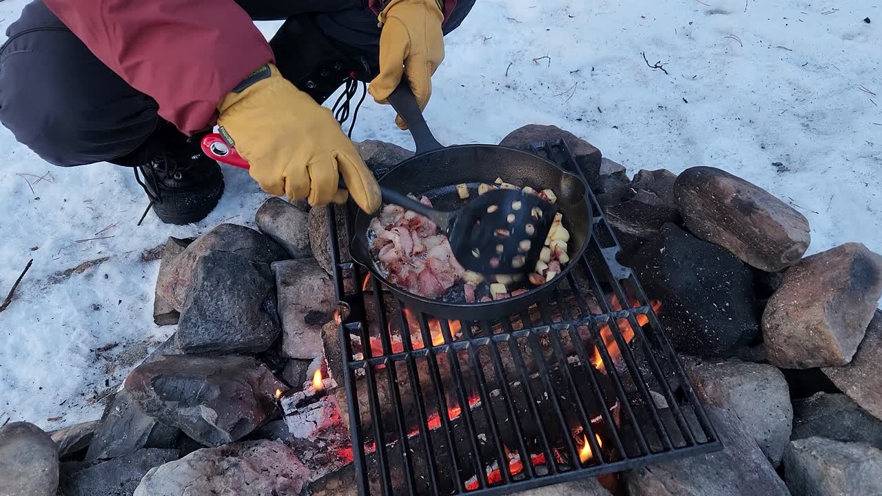 Lagning av frukost över en lägereld, vinkel 2: Rör om potatis och bacon. Tillagning av bacon och tärnad potatis i en gjutjärnspanna över en lägereld under vintercamping.