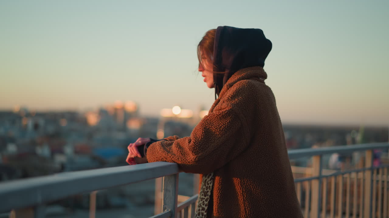 A sorrowful woman walks slowly while holding onto a metal railing, dressed in a brown coat and a hoodie, with a melancholic expression as the cityscape blurs in the background during sunset