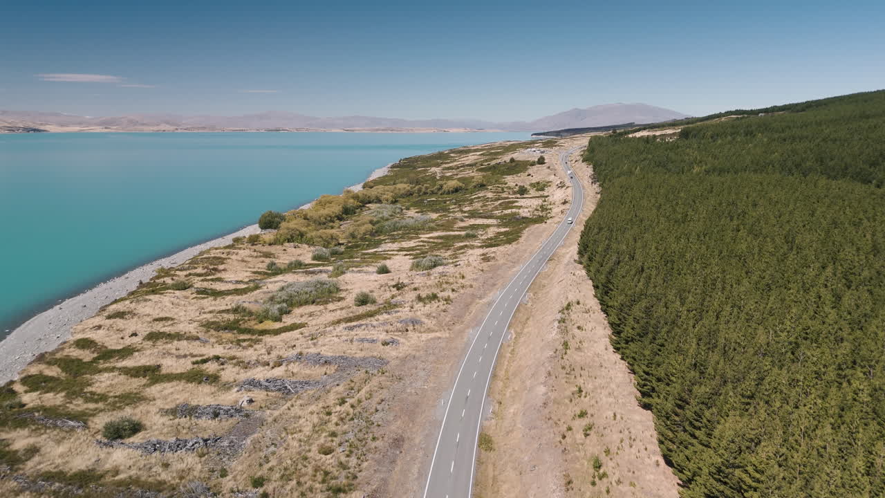 Aerial view of a road along a turquoise lake, surrounded by mountains and forest in New Zealand