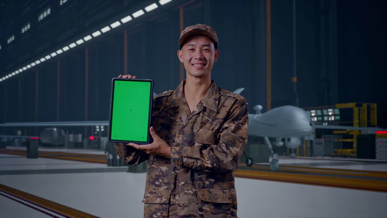 Soldado asiático sorrindo e mostrando um tablet com tela verde para a câmera, de pé com um drone militar moderno em um hangar de manutenção.