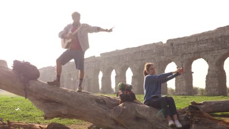 happy young couple backpackers tourists on a log trunk taking selfies photos with smartphone in front of ancient roman aqueduct ruins in romantic parco degli acquedotti park in rome misty sunrise slow motion