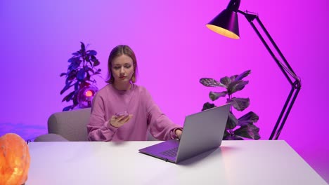 a woman in a pink sweatshirt sits at a sleek white desk, reaching for her pink smartphone while using a laptop, surrounded by plants and vibrant neon lighting in a creative home office
