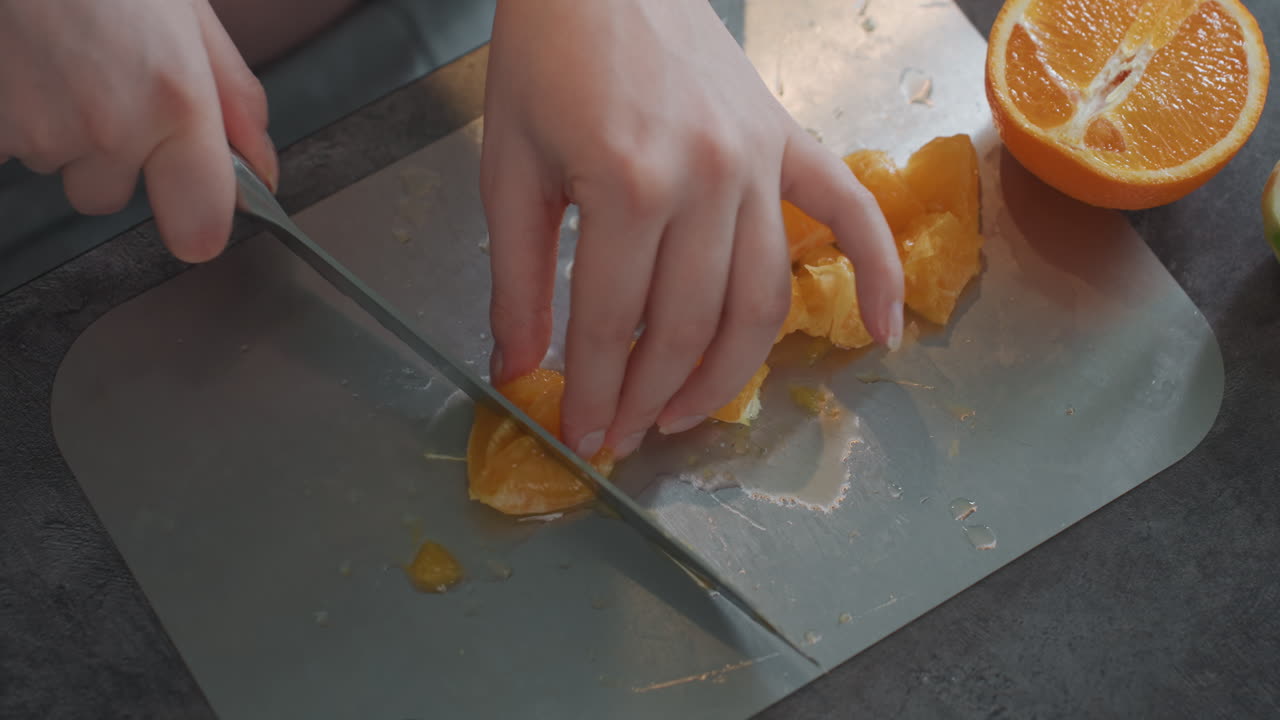 Pregnant caucasian woman slicing orange segments with steady hands near visible belly, closeup of knife and board, juice droplets and bright countertop, calm maternal routine and careful food prep