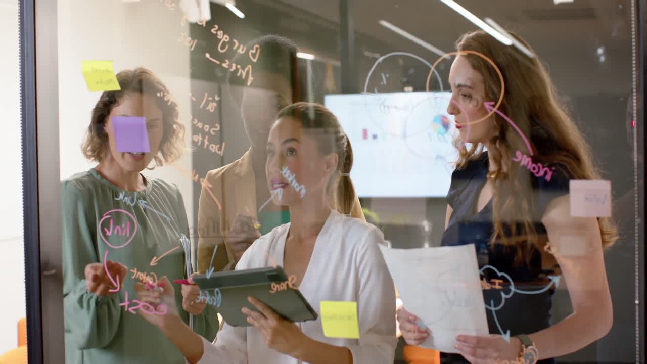 Diverse businesswomen writing on glass wall and using tablet brainstorming at office, in slow motion