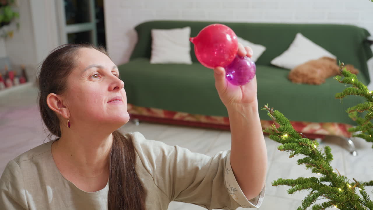 Woman smiling while lifting red and purple Christmas ornaments toward light, sitting beside decorated Christmas tree, cozy home background with green couch and festive holiday atmosphere