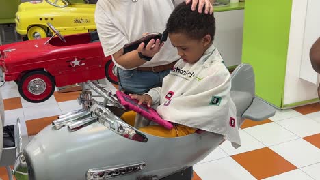 un niño negro de tres años se corta el pelo en un salón de peluquería infantil mientras su madre lo consuela.