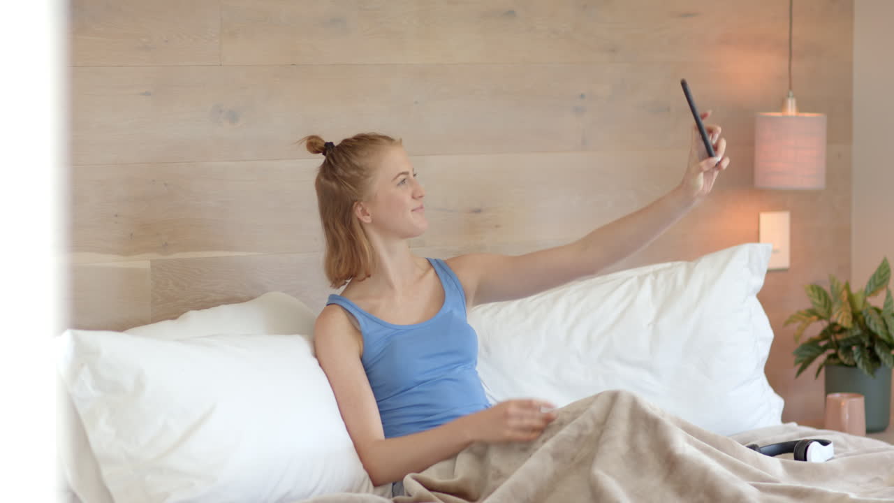 Taking selfie, woman relaxing in bed with smartphone at home