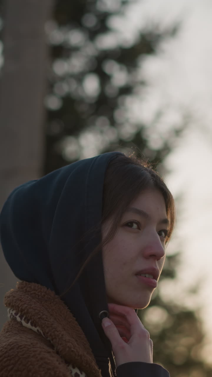 Close-up of a sad girl walking through a park at sunset, wearing a brown coat and a hoodie with the hood up, her face partially concealed, reflecting a somber mood against the soft, fading light
