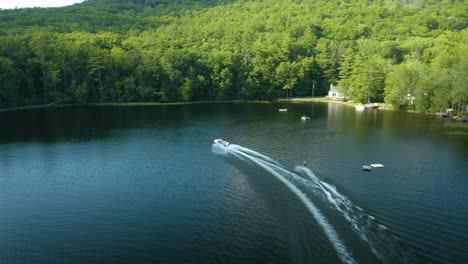 Water-skiing-dude-pulled-by-the-speedboat-in-the-outdoors-at-blue-lake-with-many-luscious,-green-trees-in-the-background-and-a-cloudy-sky,-you-can-see-splashes-of-water-or-waves