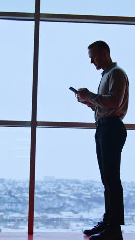 office employee at the window with phone and white cup. full-length portrait of a slim man at the backdrop of panoramic window. vertical video