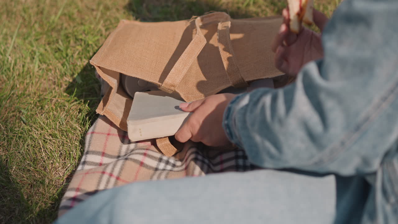 Hand holding sandwich beside picnic bag. Checkered blanket on grass, paper bag and wrapped sandwich being unwrapped book on lap, sunny afternoon, friendly casual snack moment with relaxed outdoor