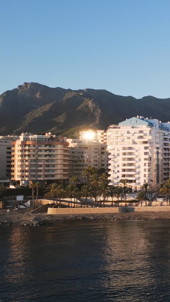 Ripresa aerea verticale con drone che inquadra la costa di Marbella durante l'ora d'oro, con il sole che si riflette sugli edifici di fronte alla spiaggia e le montagne costiere sullo sfondo