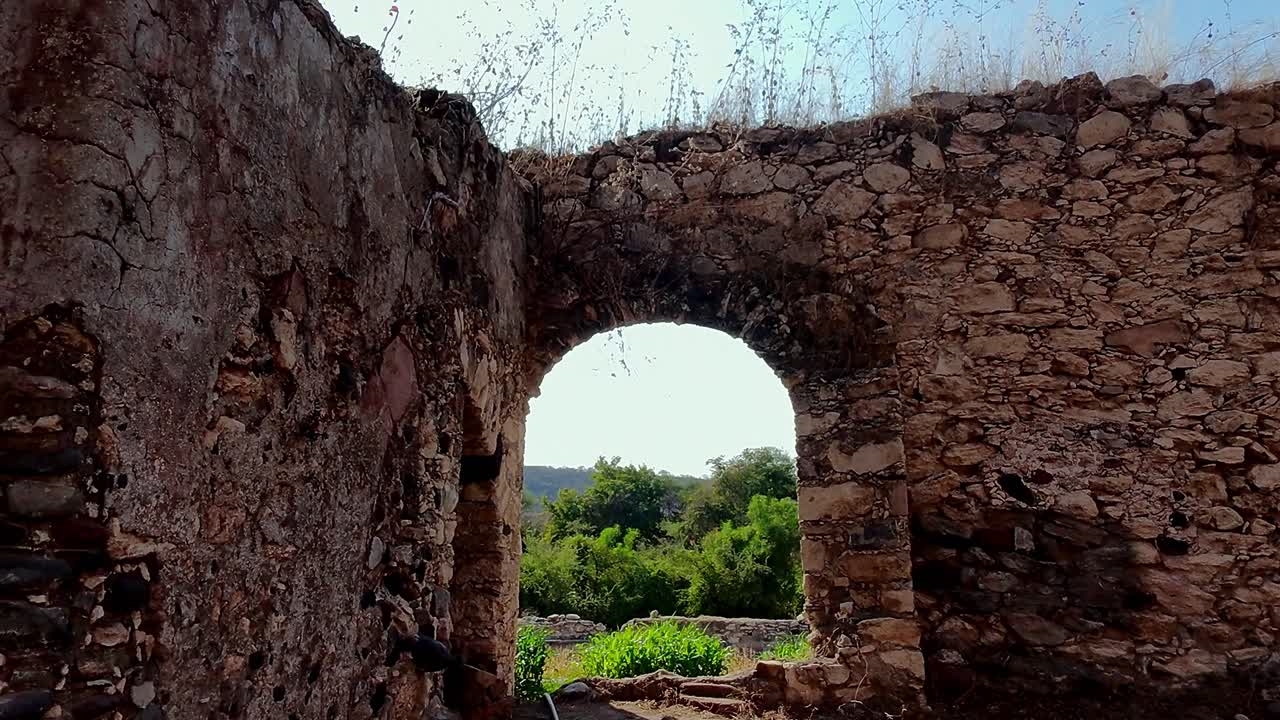 Dolly in shot through arched stone doorway revealing lush landscape, La Mezquitera, Morelos. Stone archway of old ruins frames green trees, plants, and hills under a clear blue sky