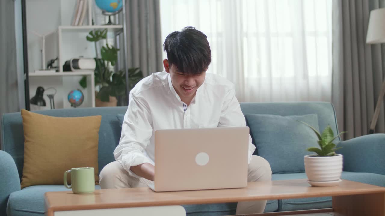 Smiling Asian Man Sitting On Sofa Use Laptop Computer In The Living Room