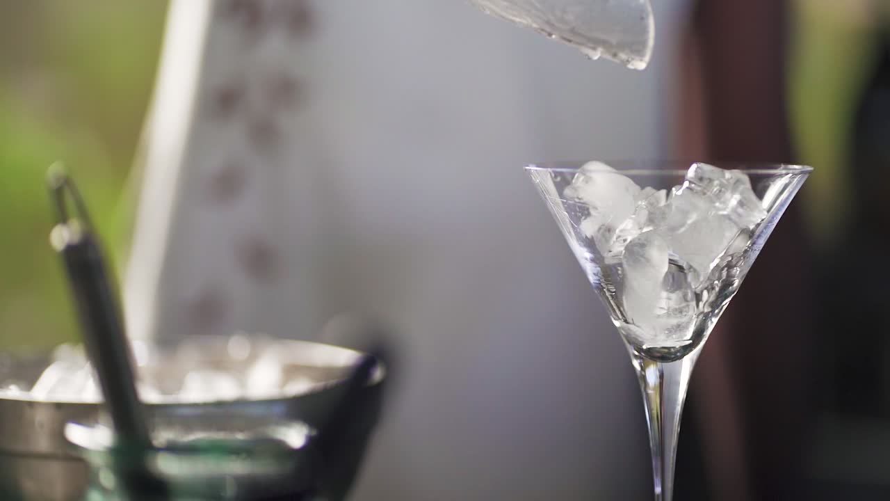 una mano vierte hielo de una cuchara en un vaso de martini en un entorno al aire libre con un cubo de hielo en el fondo