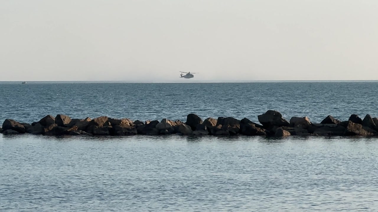 Italian Navy helicopter flying low over the sea beyond a rocky breakwater in the Gulf of La Spezia, Liguria, Italy.
