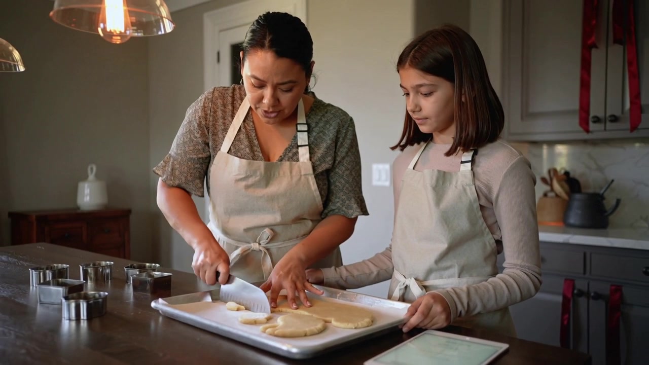 Cutting woman in aprons, placing bench knife and teaching teen at kitchen island, spacing cookies
