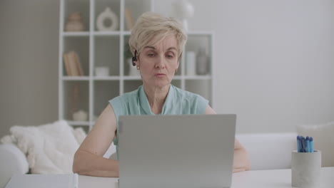 aged woman with headset is speaking by web camera using video call for communicating on laptop