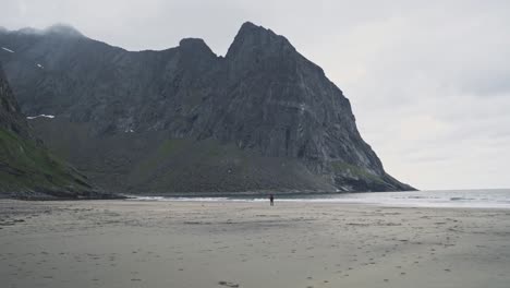 Lonely-person-walking-on-sandy-beach-near-massive-mountain-on-Norway-coastline