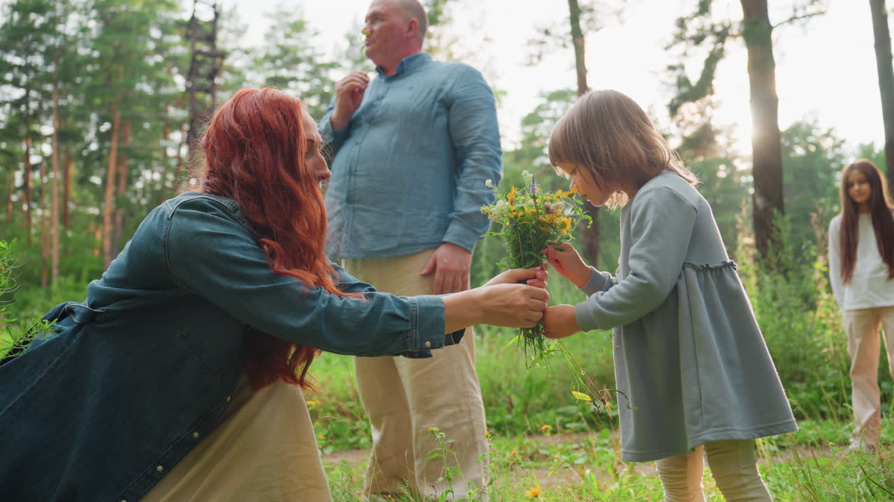 Mother hands flower to daughter to smell during peaceful walk in green forest, surrounded by sunlight and nature, capturing gentle moment of love, tenderness, and connection in family outdoor life
