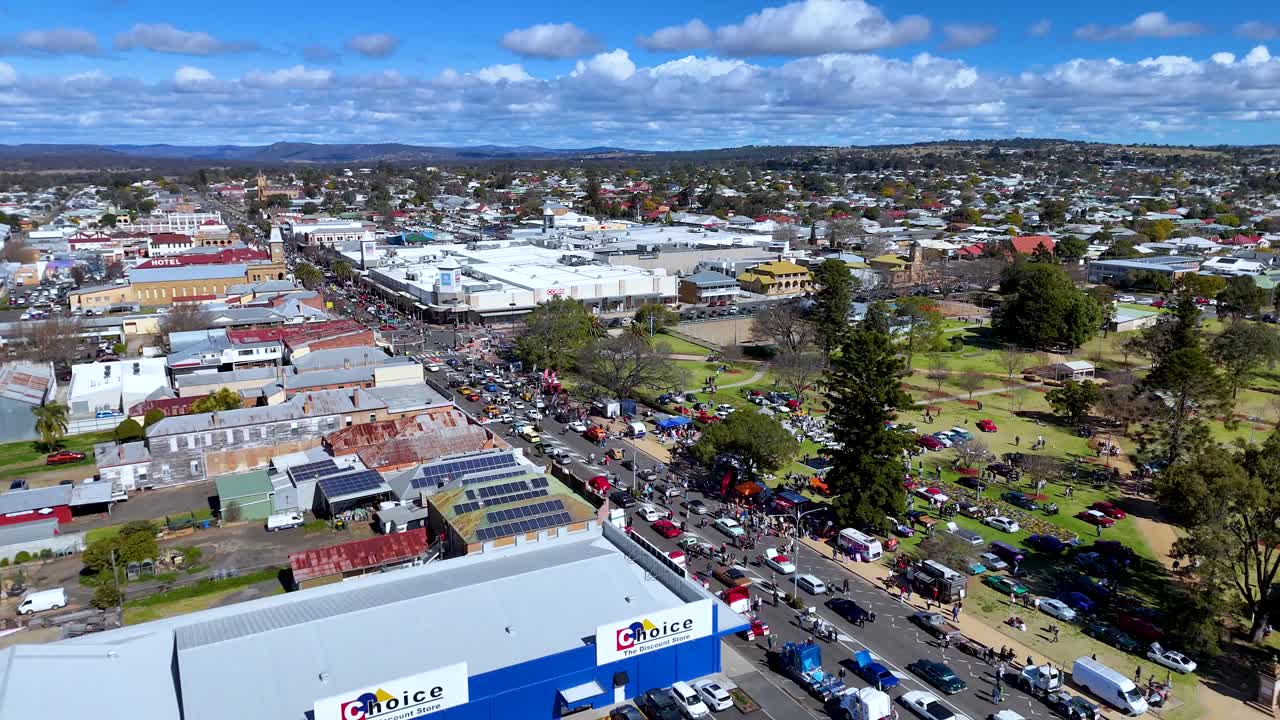 Drone footage captures a vibrant street festival in Warwick, Queensland, with crowds, parked cars, and clear blue skies. Smooth lateral camera movement reveals the bustling scene