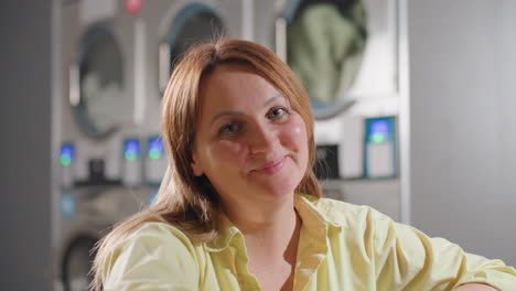 business woman seated inside laundromat opens record book, smiles with satisfaction while reading and nodding approval, cup of coffee placed on steel table with washing machines in background