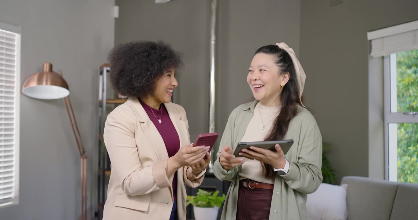 Diverse female coworkers comparing screens in living room, collaborating with pink phone and tablet