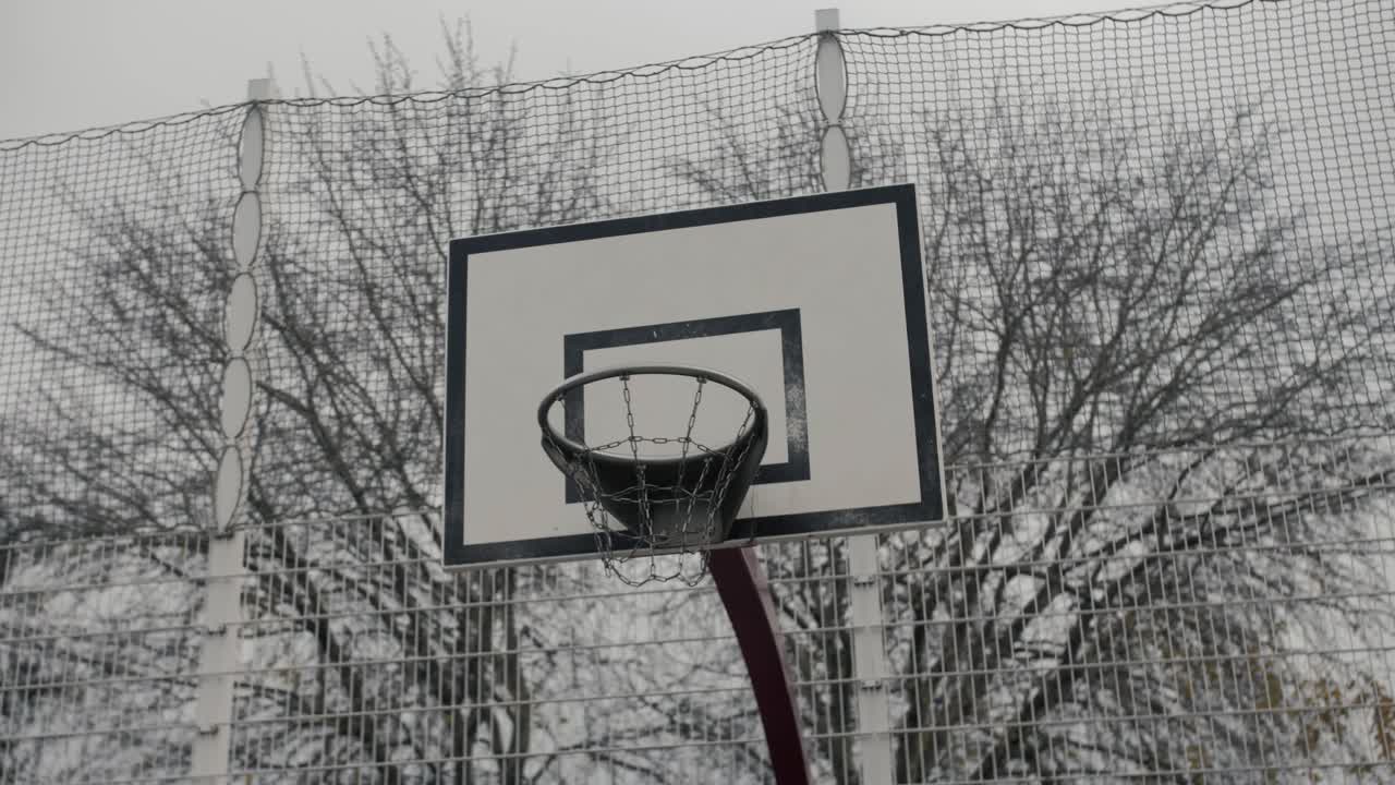 A detailed slow motion arc shot of a basketball hoop with a protective net, set against winter trees and urban surroundings