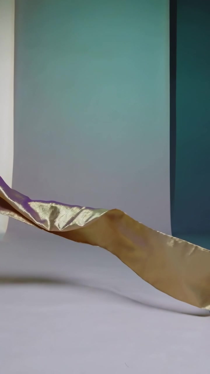 Video verticale: Fascia di raso dorato che si muove su un fondale da studio dopo essere stata tirata, mostrando luce rosa e verde acqua.