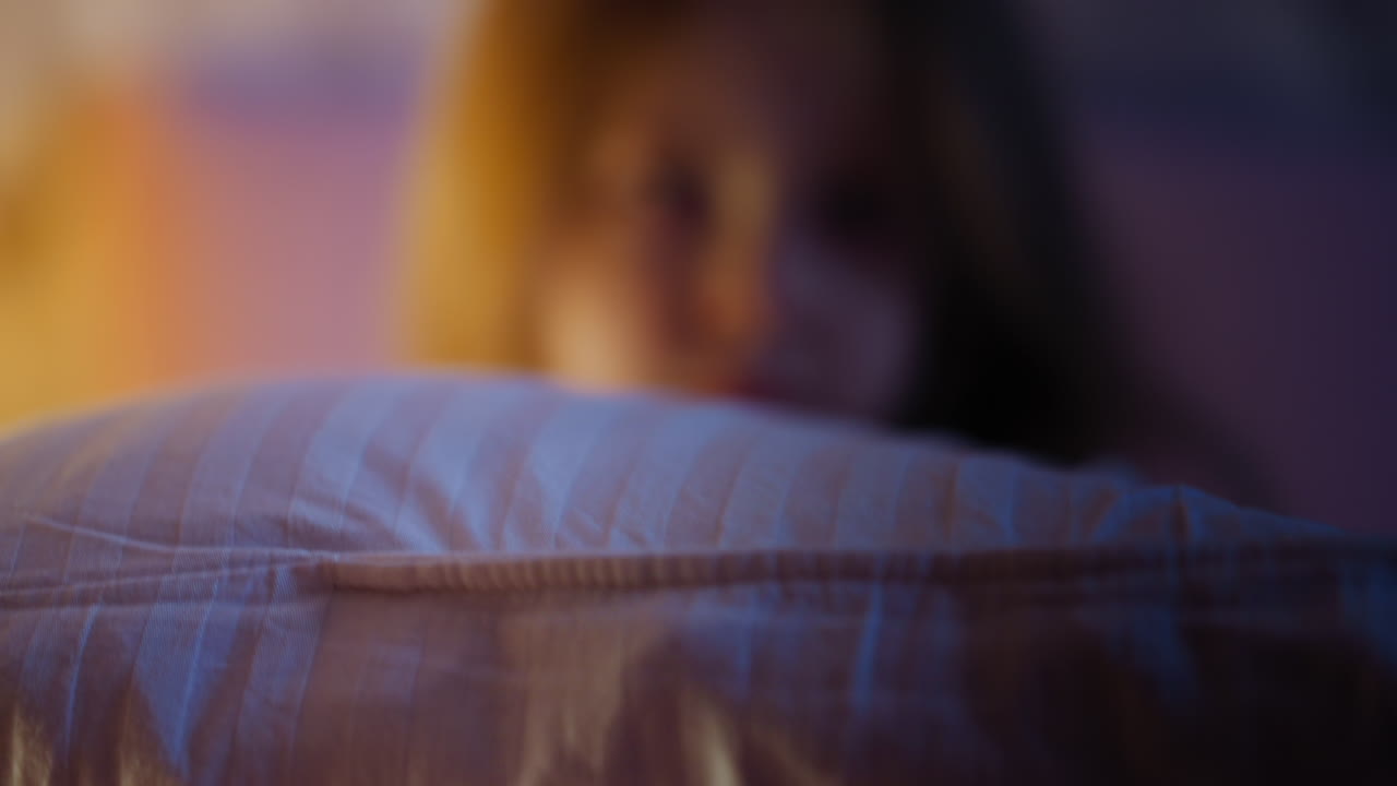 예쁜 소녀는 베개 뒤에 숨어 침실에서 시간을 보내고 있습니다.