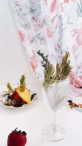 a still life with cake, strawberries, water in a wine glass, and a floral curtain.