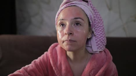 woman watching a late night movie at tv, eating popcorn. bathrobe, facial mask