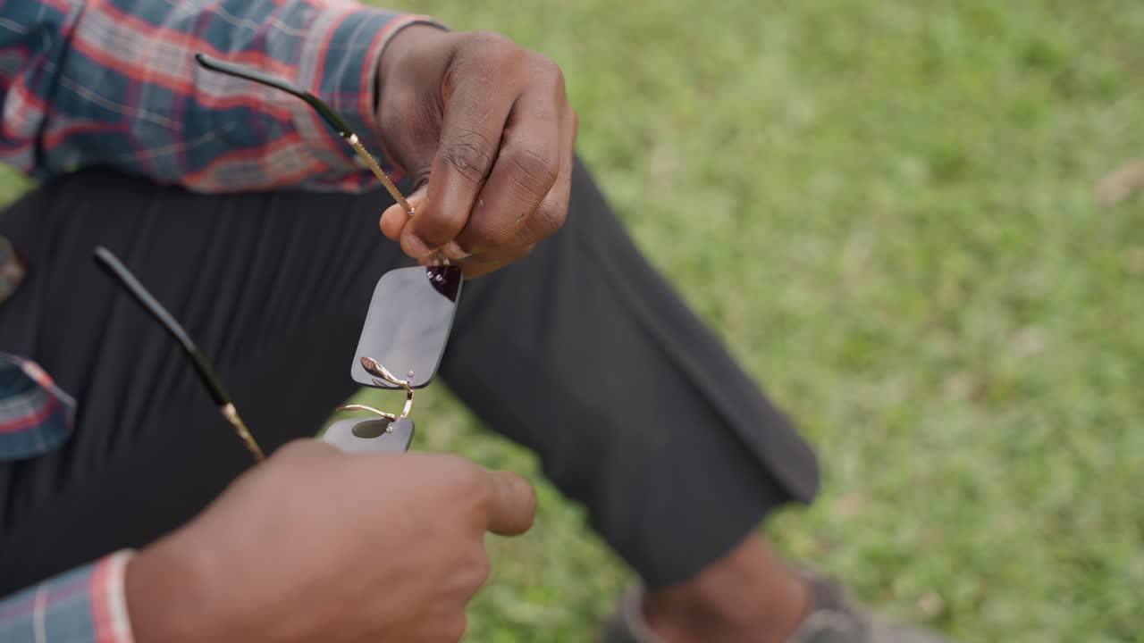 Uomo all'aperto lucida con cura una lente. Individuo rilassato in camicia a quadri pulisce con attenzione gli occhiali all'aperto. Uomo casual con abbigliamento a quadri lucida delicatamente gli occhiali su una panchina all'aperto.