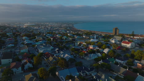 Luftaufnahme-Eines-Wohnbezirks-In-Der-Nähe-Der-Küste,-Beleuchtet-Von-Der-Untergehenden-Sonne.-Meeresbucht-Im-Hintergrund.-Port-Elisabeth,-Südafrika