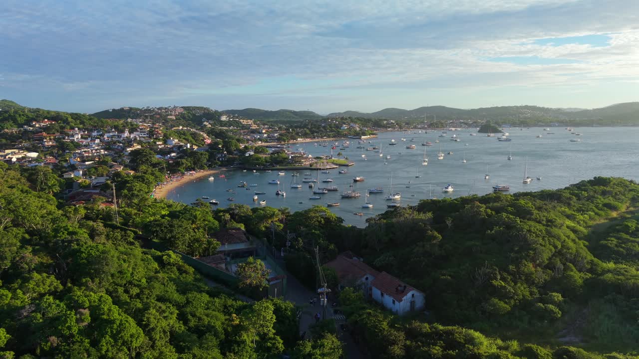 Colinas vegetadas, praia e área residencial na Praia dos Ossos, Brasil, com vários barcos atracados no oceano durante o pôr do sol. Deslizamento frontal em grande angular.