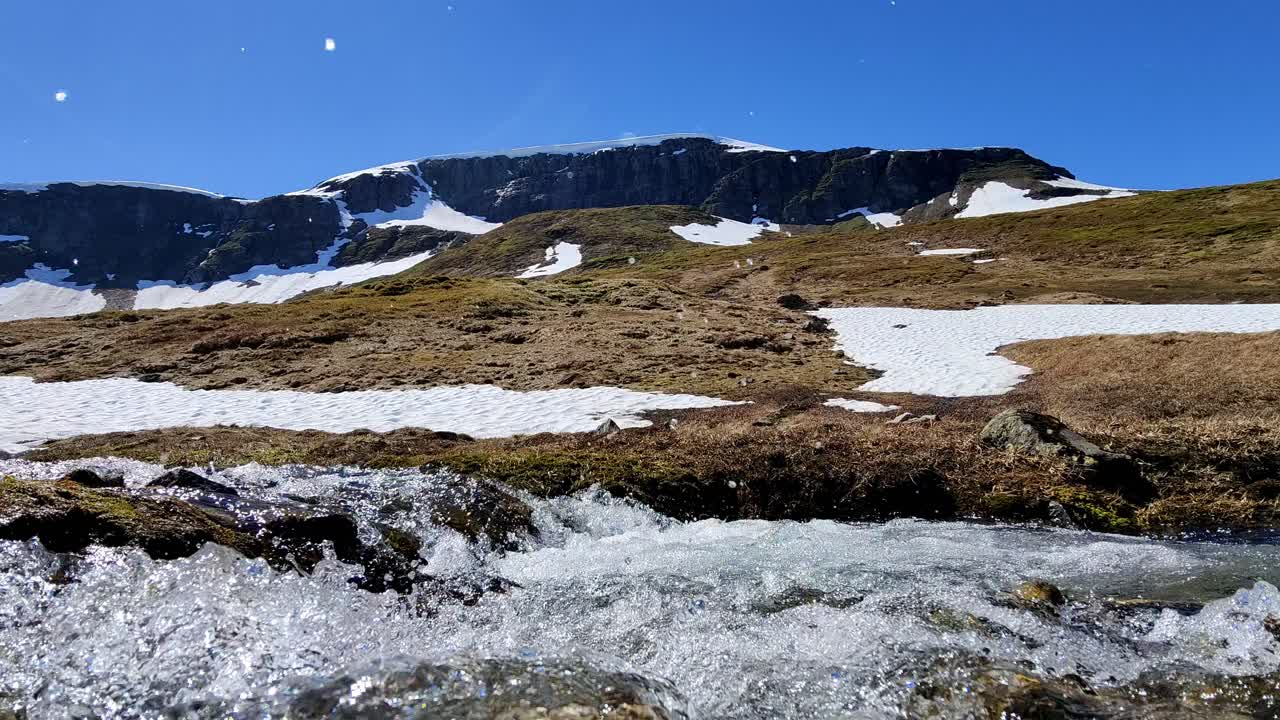 Snow melting rapidly during hot spring day and a fresh cold river is passing close in front of camera - Remaning snow cape on top of Finnbu mountain at vikafjell area in background - Static Norway