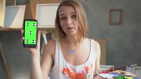 a young beautiful woman artist stands in an art studio holds her smartphone with a green screen in her hands, looks at the camera and tells something.