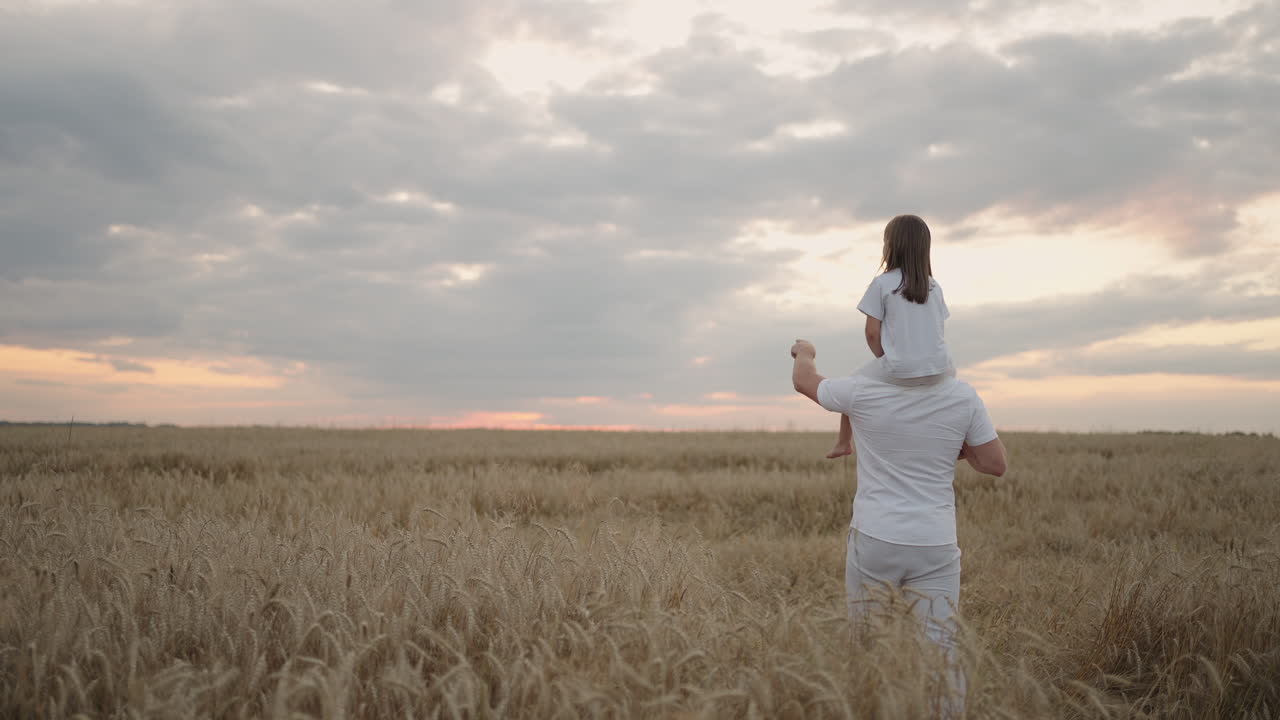 뒷면에서 볼 때: 아빠는 빛 아래에서 사랑하는 작은 건강한 딸을 어깨에 고 있습니다. 슬로모션으로 딸은 아버지와 함께 에서 걸어가고 자유롭고 행복하게 손을 높이 흔들고 있습니다.