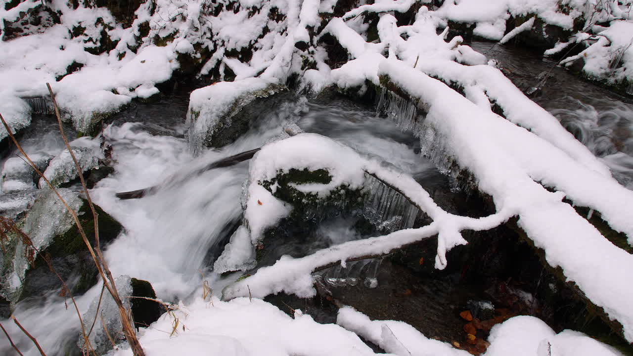 Schwenk von rechts nach links entlang eines kleinen Baches, der über Felsen und Eis in einem schneebedeckten Wald in Alaska fließt.