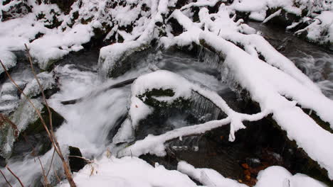 Panorámica-De-Derecha-A-Izquierda-A-Lo-Largo-De-Un-Pequeño-Arroyo-Que-Fluye-Sobre-Rocas-Y-Hielo-En-Un-Bosque-Cubierto-De-Nieve-En-Alaska