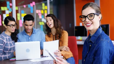 Smiling-female-executive-using-digital-tablet-while-colleagues-discussing-over-laptop